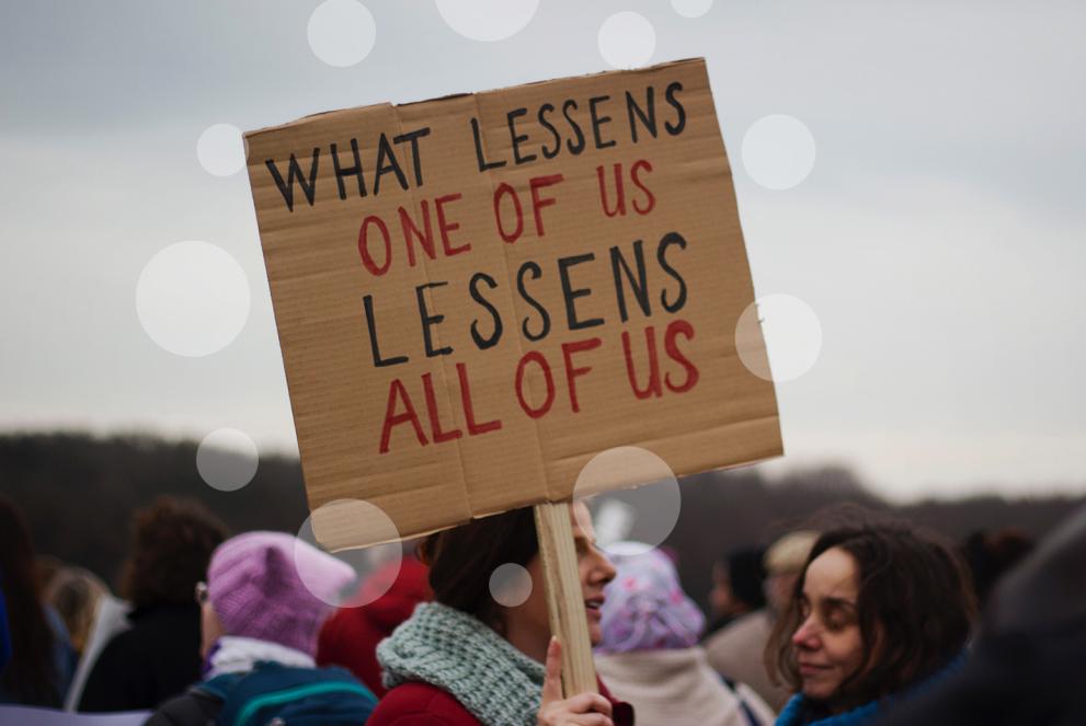 Woman holding up a sign. Text on sign: What lessens one of us lessens all of us