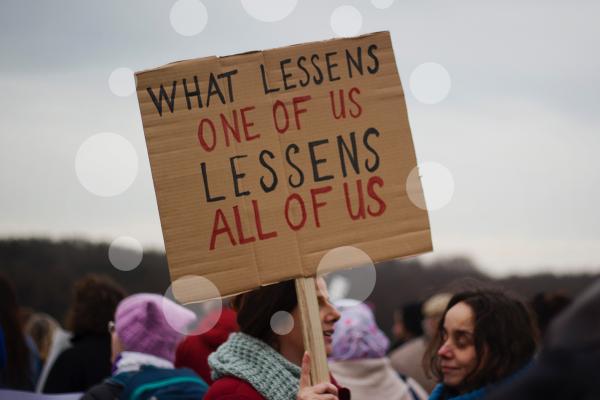Woman holding up a sign. Text on sign: What lessens one of us lessens all of us